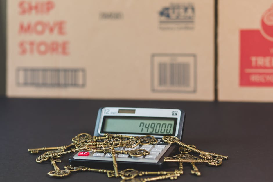 A calculator displaying the number 749800 sits on a black surface surrounded by scattered brass keys and keychains, with large cardboard boxes in the background marked with 'Ship Move Store' and barcodes. The scene suggests preparation for a home relocation involving packing and transfer of smaller items, with the keys representing household furniture or valuables to be moved. The background packaging indicates an organized moving process as part of domestic removals services offered by Removal Companies Sutton. The lighting is neutral, highlighting the details of the keys, calculator, and boxes, reflecting a typical moving-day setup focused on logistical accuracy and item handling during the furniture transport or packing stage.