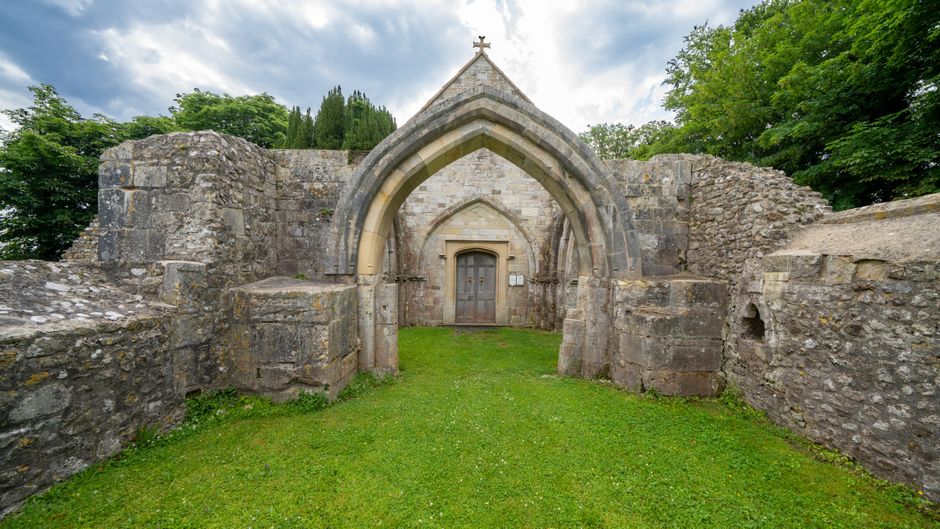 A historic stone church or chapel with a large pointed arch doorway, featuring intricate stone detailing and a wooden door, surrounded by weathered stone walls and lush green trees in the background. The entrance is set into a grassy area with well-maintained lawn, and the sky overhead is cloudy. This site appears to be an ancient or medieval structure, possibly former religious ruins or a heritage site, with the surrounding environment showing mature trees and natural greenery. The scene is well-lit with natural daylight, emphasizing the texture of the stonework and the peaceful outdoor setting. The image relates to house removals and moving services by illustrating the type of property involved in home relocation or historical building preservation projects, which Removal Companies Sutton may assist with in terms of logistics, transportation, or site clearance.