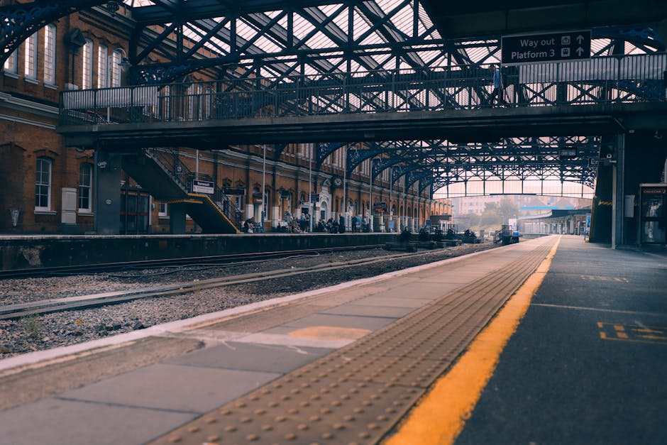 The image depicts a railway station platform with a covered metal and glass canopy overhead, supported by metal beams. The platform surface is made of concrete with a yellow tactile paving strip near the edge for safety. On the left side, a brick station building with multiple windows and staircase access is visible, alongside some waiting passengers. The railway tracks run beneath the canopy, with gravel between the sleepers. In the background, a small train or freight vehicle is seen approaching or departing, implying active rail traffic. The station appears to be well-lit with natural light suggesting daytime. This setting relates to the logistics of house removals, where transport options like train services might be considered, and occasionally [COMPANY_NAME] incorporates such elements into their detailed moving process or planning for home relocations near Sutton station, as discussed on the webpage for Sutton house removals near Sutton station. 