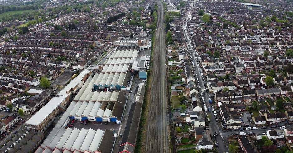 Aerial view of a residential neighbourhood with densely packed terraced houses, alongside a large industrial warehouse complex with multiple metal-roofed buildings, situated next to a railway line that runs vertically through the centre of the image. The photograph shows the exterior of a house in the process of home relocation, with visible cardboard boxes and packing materials inside and outside the property, near the entrance and on the pavement. A removal company vehicle is parked nearby, and various furniture pieces, wrapped in blankets and plastic wrap, are either being prepared for loading or are already in transit. The scene captures the loading process typical of furniture transport and house removals, with equipment such as trolleys possibly used for moving large items across the property’s entryway and onto the van. Natural daylight illuminates the area, highlighting the organized setup involved in a professional move, consistent with services offered by Removal Companies Sutton for local house removals near Sutton station, emphasizing logistics, packing, and transport procedures.