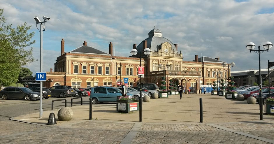The image depicts a railway station platform with a covered metal and glass canopy overhead, supported by metal beams. The platform surface is made of concrete with a yellow tactile paving strip near the edge for safety. On the left side, a brick station building with multiple windows and staircase access is visible, alongside some waiting passengers. The railway tracks run beneath the canopy, with gravel between the sleepers. In the background, a small train or freight vehicle is seen approaching or departing, implying active rail traffic. The station appears to be well-lit with natural light suggesting daytime. This setting relates to the logistics of house removals, where transport options like train services might be considered, and occasionally [COMPANY_NAME] incorporates such elements into their detailed moving process or planning for home relocations near Sutton station, as discussed on the webpage for Sutton house removals near Sutton station. 